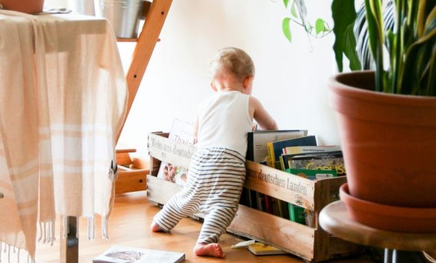 A baby standing, leaning against a crate of books